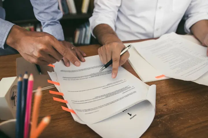 Twee personen bekijken en bespreken een contract aan een houten tafel, waarbij één persoon met een pen op de tekst wijst en meerdere pagina’s met markeerstrookjes zichtbaar zijn.