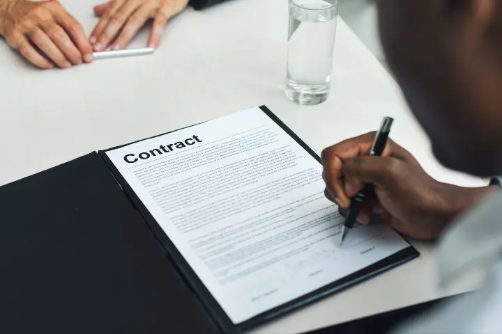 Persoon tekent een contract terwijl een andere persoon tegenover zit tijdens een zakelijke afspraak.